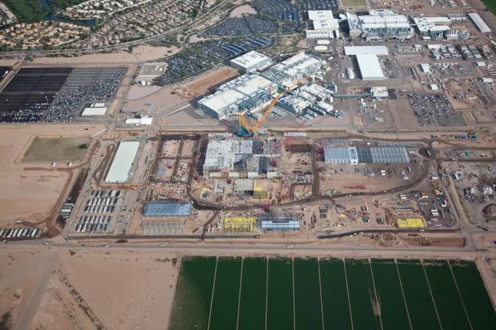 Aerial view of a large industrial construction site with cranes, surrounded by fields and parking lots.