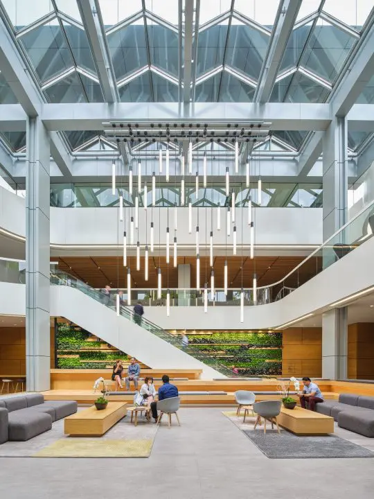 Modern atrium with skylights, seating areas, and a green wall. People are socializing and working in a bright, open space.