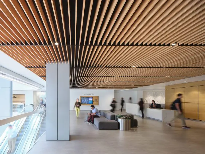 Modern lobby with wooden ceiling, people walking, sitting area, and informational displays. Bright, spacious, and welcoming environment.