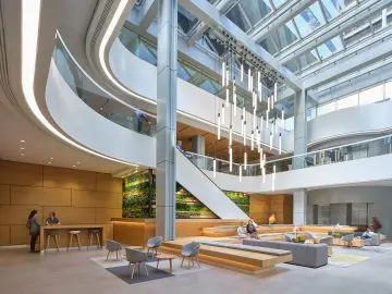 Modern office atrium with seating, greenery, and modern lighting. People interacting and working in a spacious, bright environment.