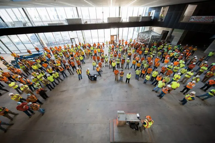 Large group of construction workers in safety vests gather inside a building for a meeting, led by a speaker on a platform.