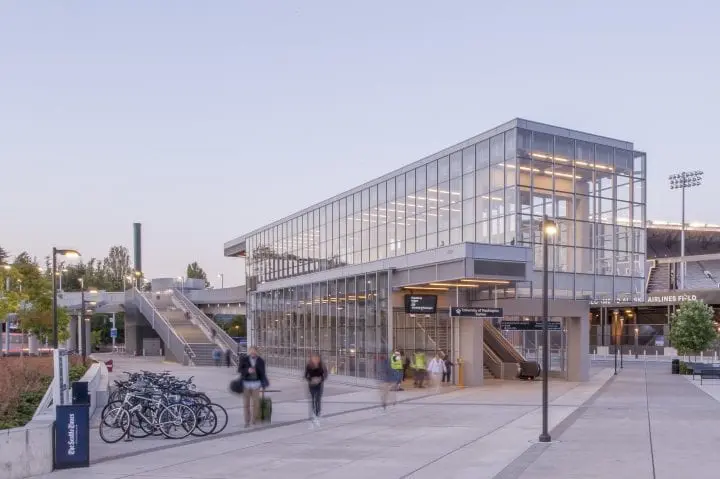 Modern transit station with glass facade, people walking, bicycles parked nearby, and steps leading up.