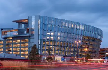Modern glass office building at dusk, illuminated against a cloudy sky. Busy street with light trails in foreground.