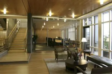 Modern office lobby with wooden floors, stairs, glass-walled conference room, and cozy seating. Bright lighting and large windows.