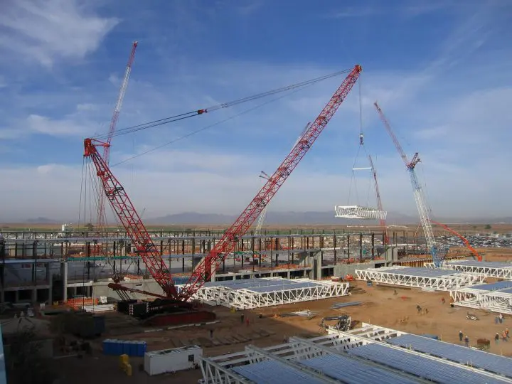 Cranes at a construction site lift steel beams, with mountains in the distance under a clear blue sky.