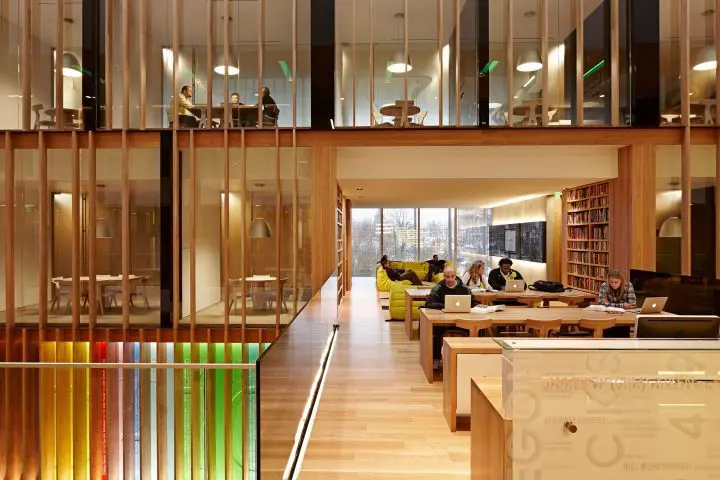 Modern library interior with people studying on laptops, wooden decor, colorful accents, and glass-walled meeting rooms above.