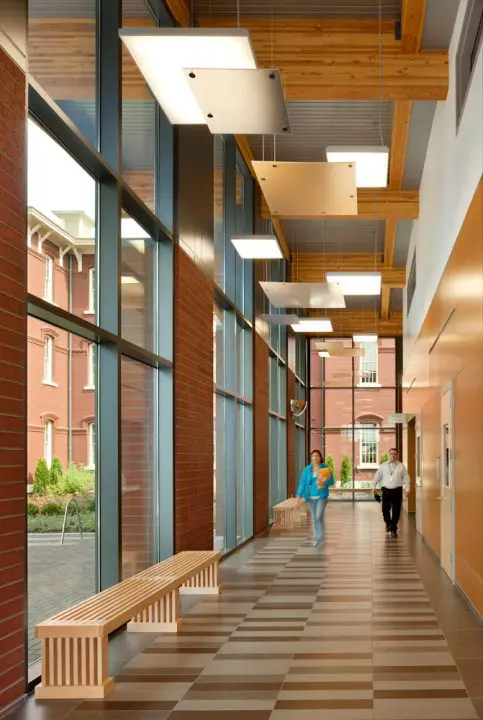 Spacious modern hallway with wooden benches and ceiling lights, featuring two people walking by large windows and brick walls.
