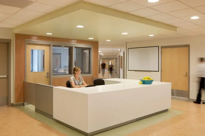 Reception desk in a bright, modern hallway with staff member seated, surrounded by wooden doors and soft lighting.