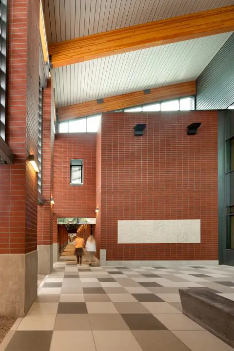 Modern architectural hallway with red brick walls, wooden beams, and tile floor. Two people walk under the checkered ceiling.
