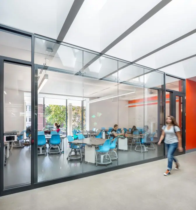 Modern classroom with glass walls, students engaged, and a person walking by in a bright, open educational space.