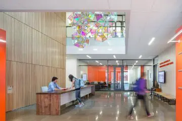 Modern lobby with colorful geometric ceiling art and people interacting at a wooden reception desk, bright and spacious atmosphere.