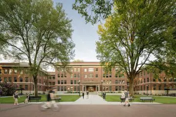 Historic red-brick university building with lush trees, students walking and cycling on the path, sunny day.