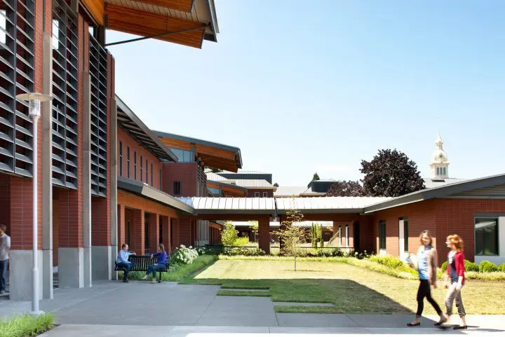 Modern campus courtyard with people walking and sitting, featuring brick buildings, greenery, and a clock tower in the background.