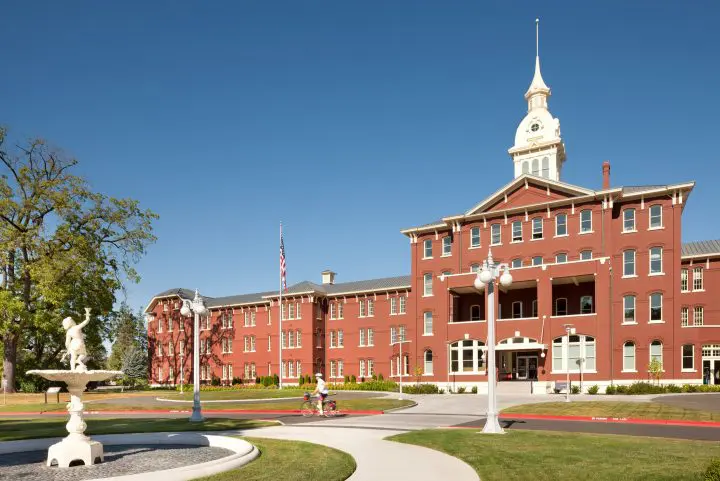 Historic red-brick building with a central tower, fountain, and clear blue sky. Person biking by, USA flag flying. Classic architecture.