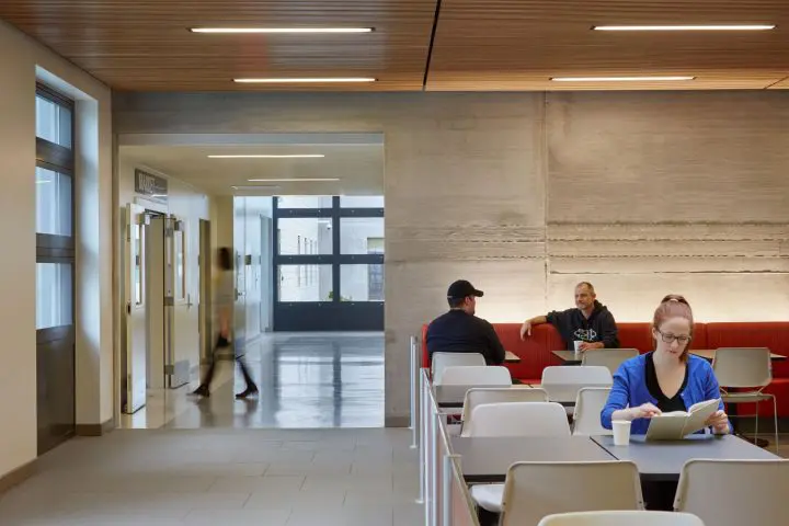 Modern lounge with people enjoying conversation and reading. Sleek design, red seating, concrete walls, and soft lighting.
