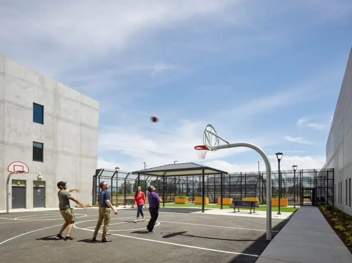 People playing basketball on an outdoor court under a clear sky, surrounded by modern urban structures.