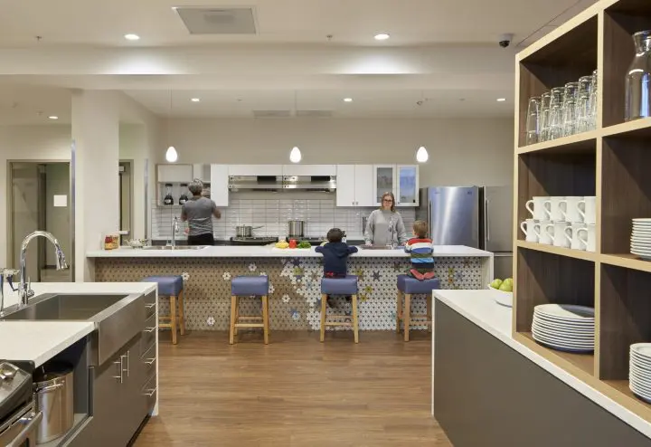 Modern kitchen interior with people cooking and interacting at a stylish island, featuring open shelves with cups and plates.