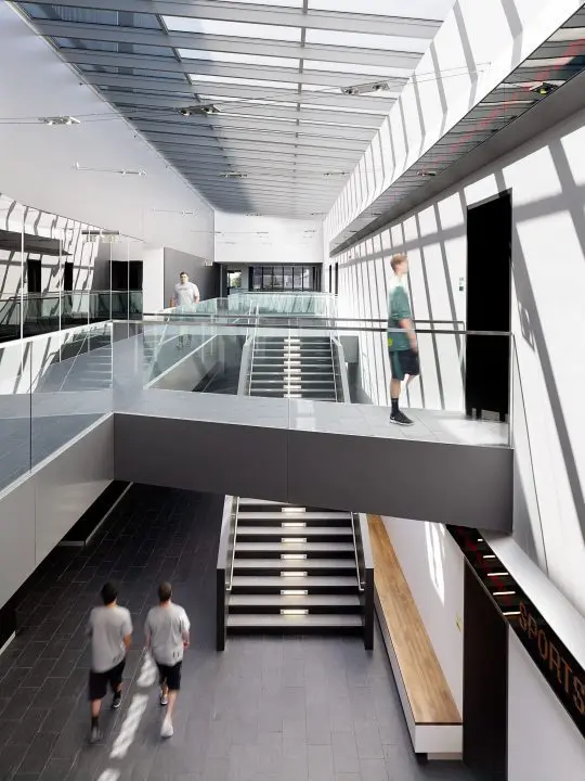 Modern atrium with people walking across a skybridge and stairs. Natural light fills the spacious architectural interior.