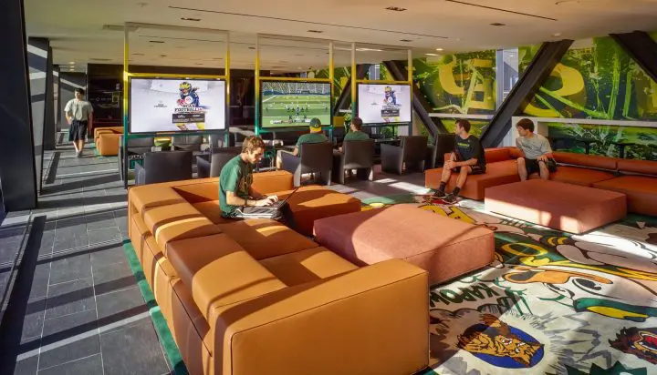 Modern gaming lounge with multiple screens, people engaged on couches, colorful decor, and natural light streaming in.