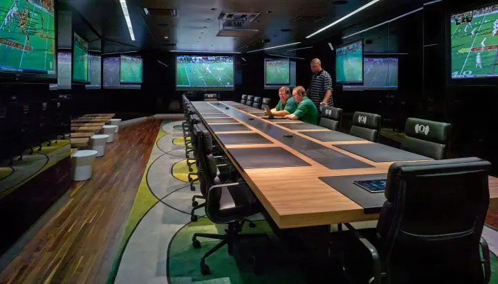 Modern conference room with attendees reviewing sports footage on multiple screens. Large table, sleek chairs, and wood flooring.