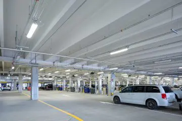 Modern parking garage interior with multiple cars parked, lit by overhead fluorescent lights, featuring concrete columns and ductwork.