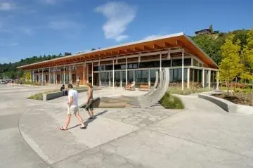 Modern waterfront building with people walking, surrounded by greenery and architecture.