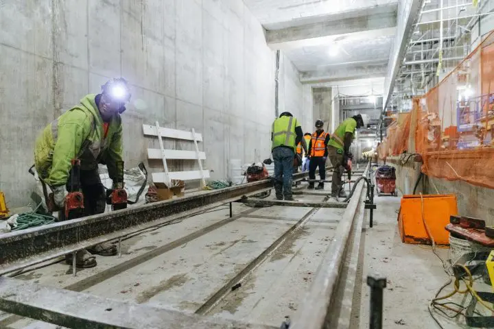 Construction crew installing underground railway tracks in a spacious tunnel, wearing safety gear and high-visibility vests.