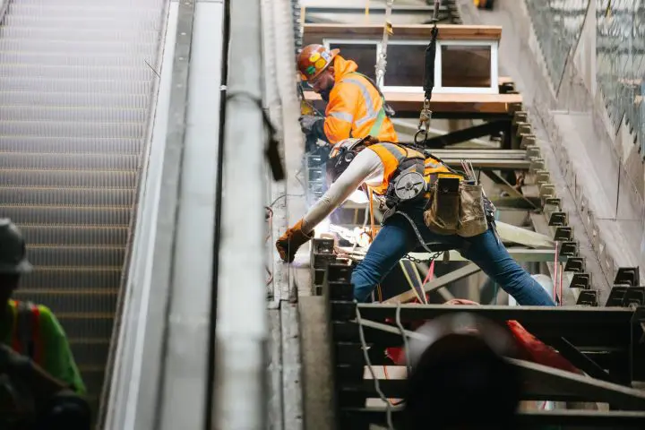Construction workers operating on escalator maintenance, harnessed for safety and wearing high-visibility gear.
