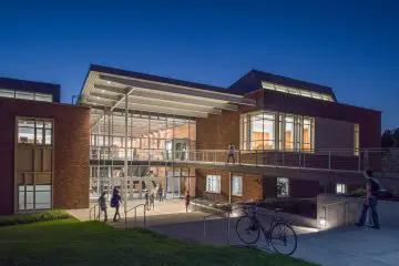 Modern building with large glass windows and walkway, illuminated at night. People walking towards the entrance, bicycle parked nearby.