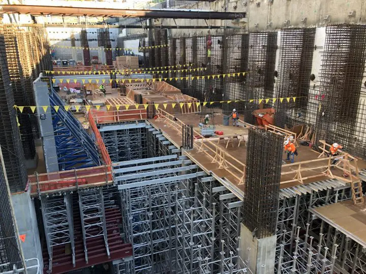 Construction site with workers on scaffolding and metal frames, building infrastructure. Safety gear and yellow flags visible.