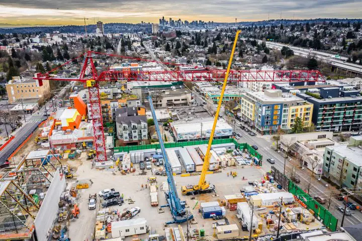 Urban construction site with cranes and buildings under a cloudy sky, city skyline visible in the background.