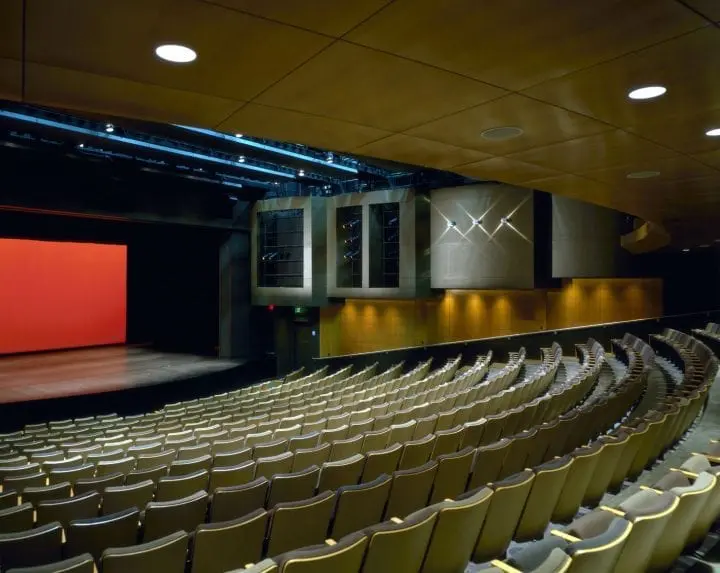 Spacious auditorium with empty rows of seats facing a stage with a red backdrop, modern lighting, and wooden interior design.