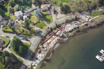 Aerial view of a lakeside construction site near residential area, showcasing progress and surrounding greenery, water, and roads.