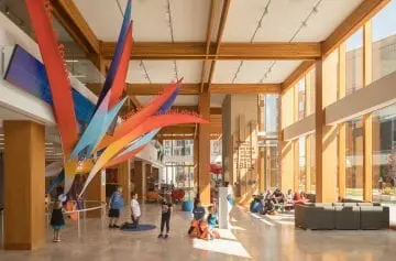 Bright, spacious lobby with large wooden beams and colorful art installation, featuring people interacting and relaxing in natural light.