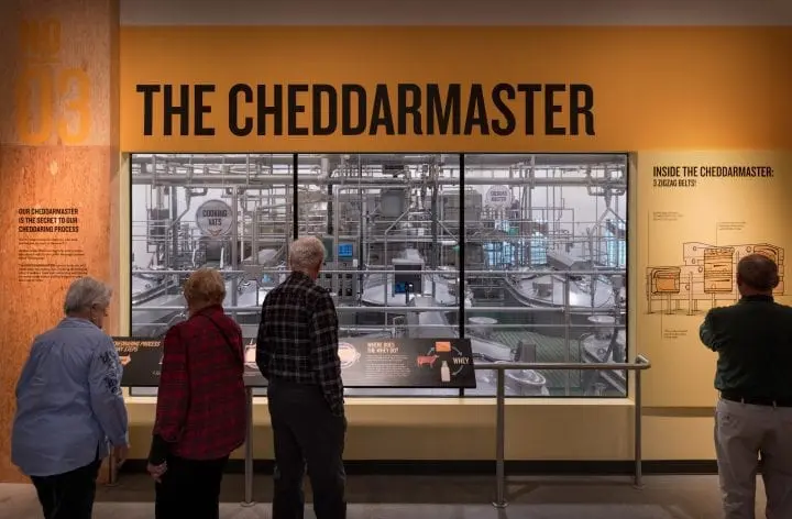 Visitors observe cheese-making process at The Cheddarmaster exhibit, showcasing industrial equipment behind large viewing windows.