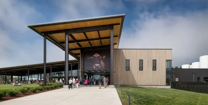 Modern building with large cow image on facade, visitors walking towards entrance. Outdoor seating on grassy area, under a clear sky.