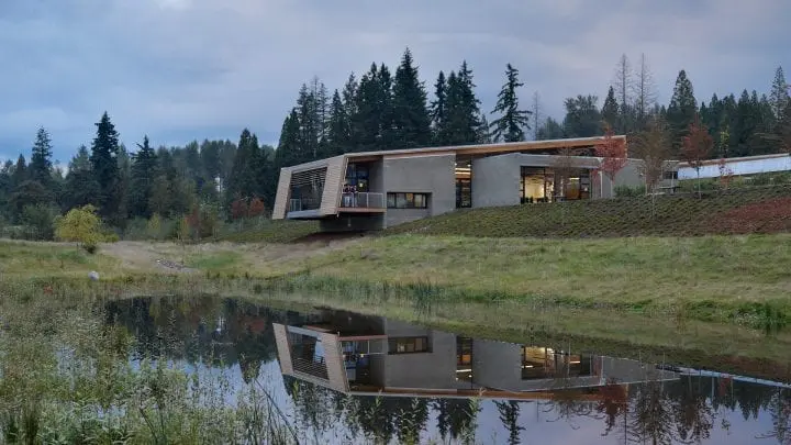 Modern building with large windows set in a serene, wooded landscape reflecting in a calm pond under a cloudy sky.