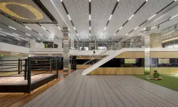 Modern gym interior with boxing ring, a person stretching on artificial turf, and another walking upstairs in a sleek, bright setting.