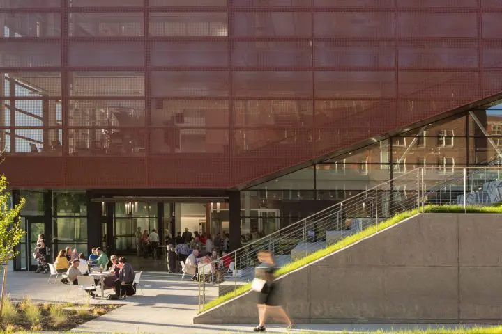 Modern building with glass facade; people socializing outside, green space and stairs in view. Sunny day.