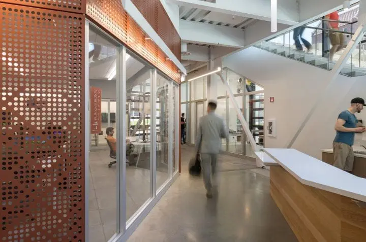 Modern office space with individuals working, ascending stairs, and engaging in tasks. Open design featuring glass walls and orange accents.