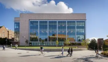Modern university building with glass facade, students walking and biking. Blue sky and green lawn in the background.