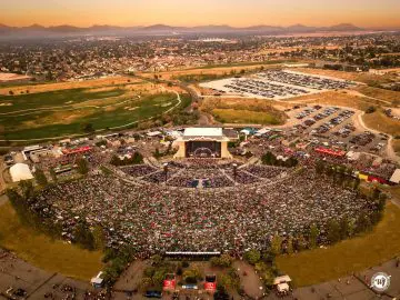 Aerial view of a large outdoor concert at sunset, with a packed crowd, stage, and surrounding city landscape.
