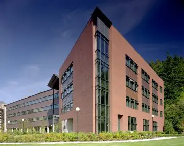 Modern brick office building with large windows, surrounded by greenery under a clear blue sky.