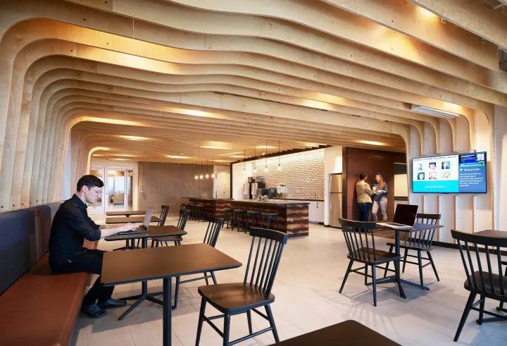 Modern office lounge with wooden ceiling, tables, and seating. Person working on laptop, two people chatting near coffee area.