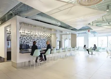 Modern office kitchen with people collaborating at bar and tables, bright interior design, pendant lighting, and checkerboard tiles.