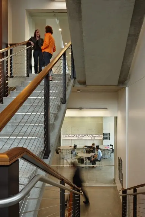 A modern staircase with two people talking, others collaborate in a glass-walled room below. Bright, open office environment.