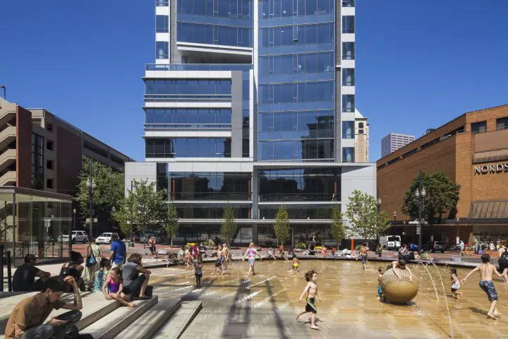 Urban plaza with people enjoying a sunny day near fountains, tall glass building, and Nordstrom store in background.