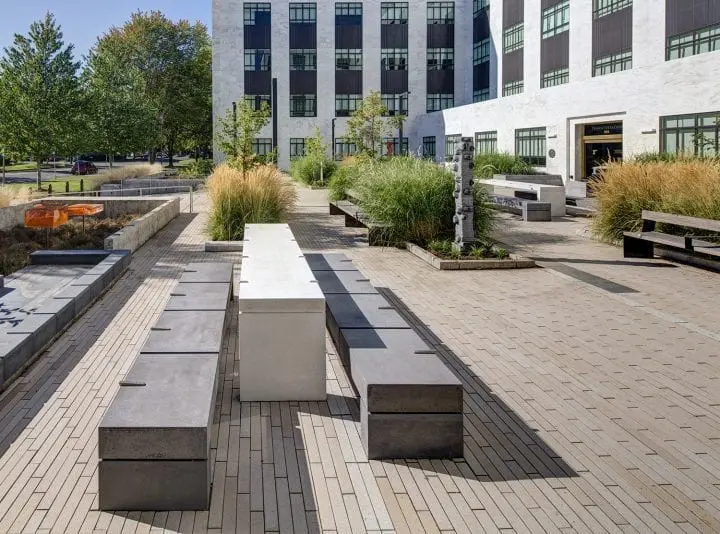Modern urban courtyard with sleek benches, lush landscaping, and tall building backdrop, under clear blue sky.
