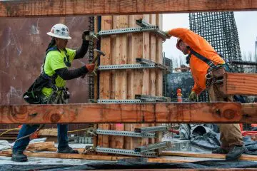 Construction workers reinforcing a wooden column with metal brackets on a job site. Safety gear and teamwork in action.
