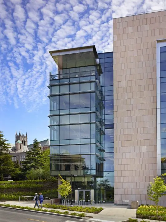 Modern glass building exterior with two people walking by, under a cloudy blue sky. Urban architecture and greenery.